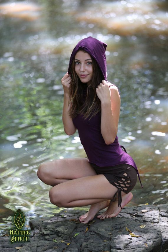 a young woman in a purple hooded tank top and brown shorts sits by a river. she is smiling and looking at the camera. she is surrounded by beautiful natural scenery.  the photo has a bohemian/hippie style.