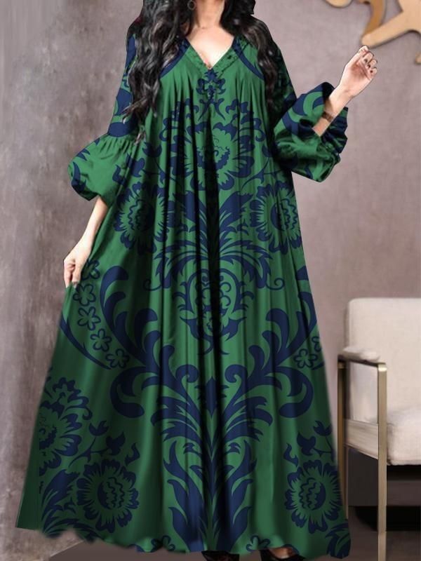 an image shows a woman wearing a stunning green maxi dress with a vneck and long, voluminous puff sleeves. the dress features a beautiful navy blue damask floral print and a pleated design. the dress has a loose and flowing fit, perfect for various occasions.  the woman has long dark hair and is posing against a muted backdrop. a portion of a chair is visible in the background.