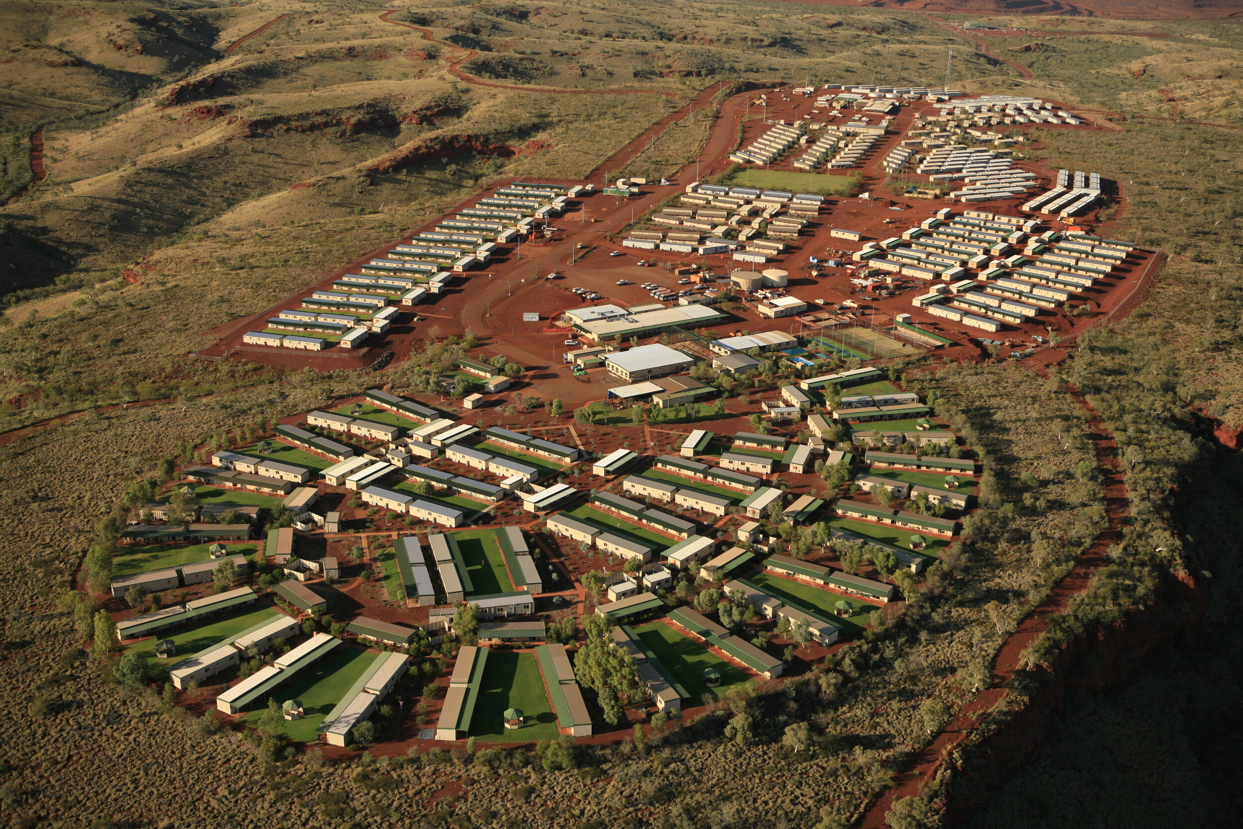 an aerial view of a mining camp in the australian outback. the camp is situated on a red dirt plain, surrounded by sparse vegetation. the camp is made up of a large number of buildings, including housing for workers, offices, and other facilities. the camp is remote and isolated, and the workers who live there are required to fly in and out. the camp is an important part of the mining industry in australia, and it provides a place for workers to live and work while they are away from their families.