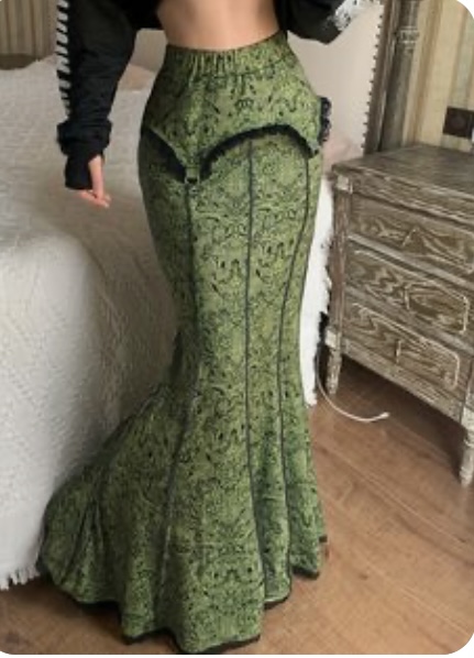 a woman is standing in front of a bed and a dresser, wearing a long green skirt with a lace trim. the skirt is fitted at the waist and hips, and flares out into a mermaidstyle hem. the woman is also wearing a black longsleeved top. the room is welllit with natural light.