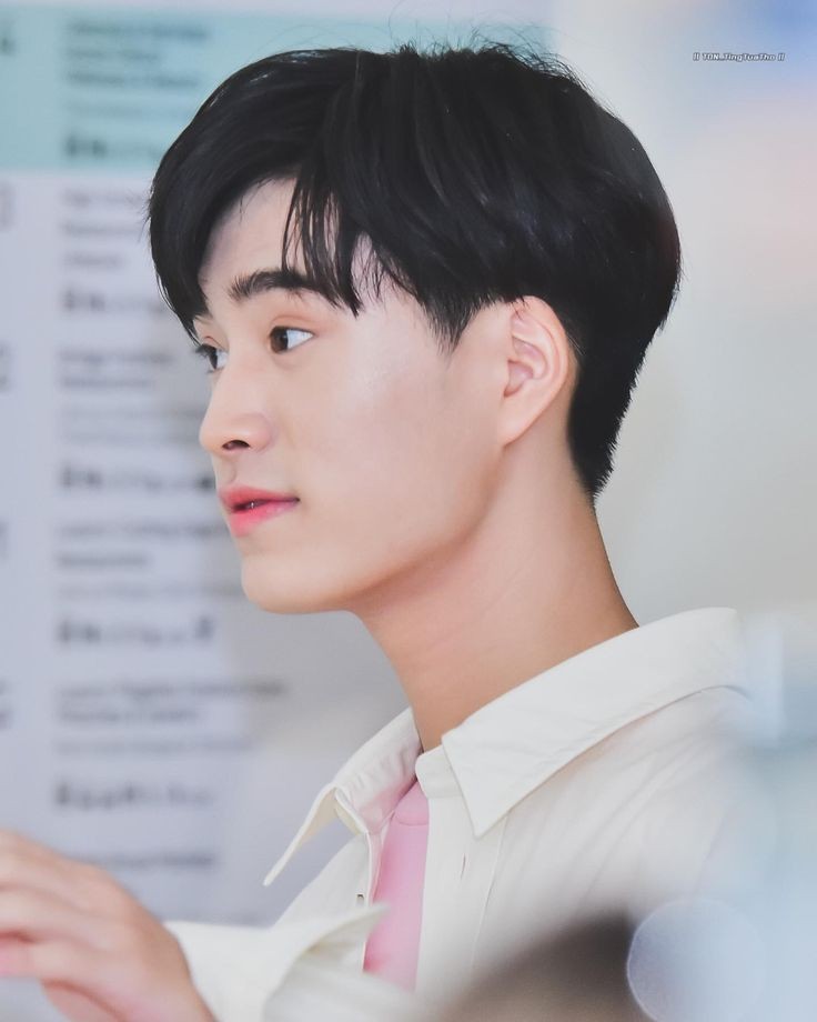 a head and shoulders side profile photo of a young asian man wearing a white, collared shirt over a pink shirt. the background is blurry and out of focus.