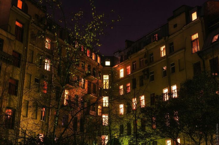 the image shows a nighttime view of two buildings forming a courtyard. the buildings are old, multistoried, with many windows lit from inside, casting a warm orange glow. the buildings are a tan or yellowish color, and the windows are rectangular.  a tree with bare branches partially obscures the view, adding a layer of depth and mystery. the overall atmosphere is dark, moody, and atmospheric.  the lighting focuses on the illuminated windows, drawing attention to the residents within the buildings and creating a sense of life within the seemingly quiet urban space.