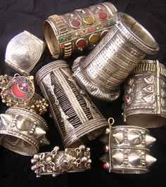 a pile of assorted silver bangles with intricate designs. some are adorned with semiprecious gemstones and others feature embossed and detailed artwork. they're piled on a dark surface and are reminiscent of traditional indian craftsmanship.