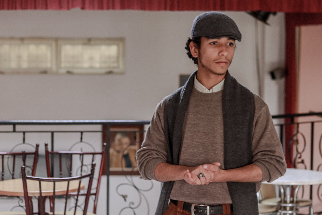 the image is a medium shot of a young man standing indoors, possibly in a cafe or restaurant. he is wearing a brown turtleneck sweater, a grey scarf, and a newsboy cap, giving him a vintage or retro 1920s/30s style. his expression is thoughtful or pensive. the background features wooden tables and chairs in a cafelike setting, with a railing suggesting a balcony or raised area.  the overall setting and his attire evoke a sense of style and elegance.