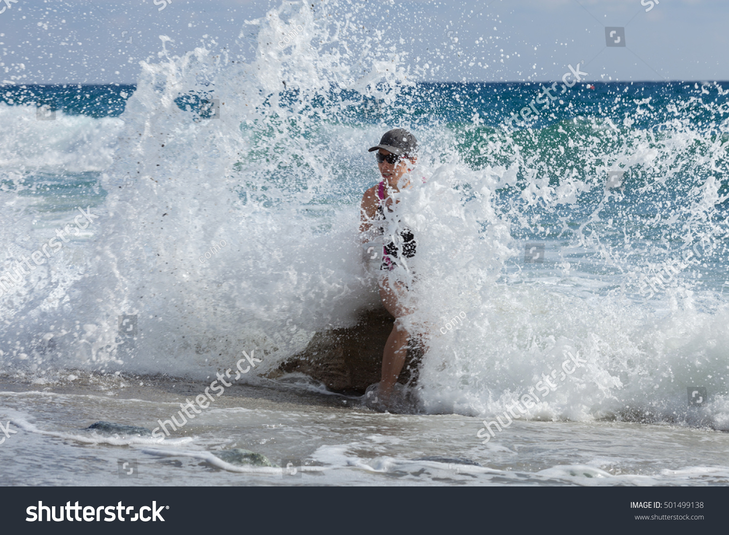 a woman in a swimsuit and a baseball cap stands in the ocean as a large wave crashes over her. the beach is visible in the foreground and the ocean extends to the horizon.