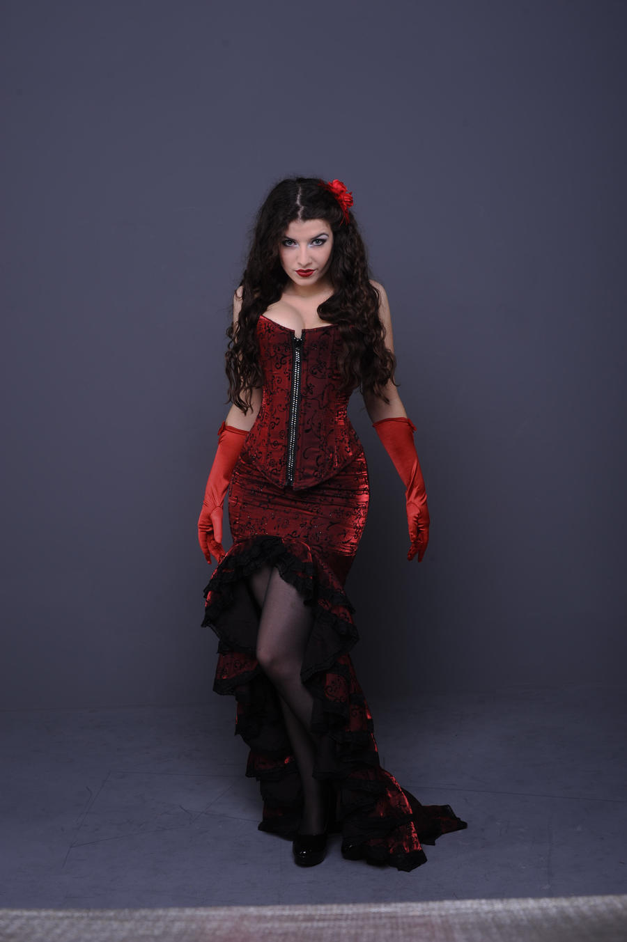 a full shot of a woman wearing a long red corset dress and red gloves. she has dark curly hair and is posing confidently against a gray background.  a single red flower is in her hair. she appears to be in a studio setting and the overall style is evocative of vintage glamour or gothic fashion.