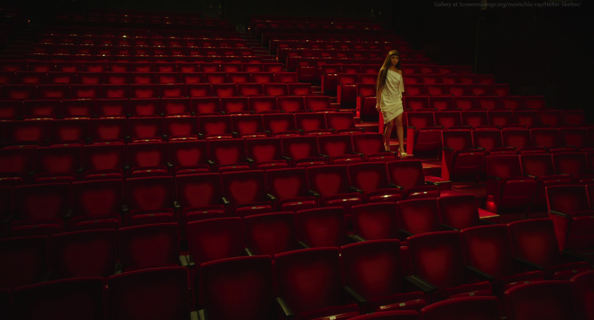 a woman in a white dress walks down the aisle of an empty theater with red seats. the lighting is dim and dramatic.