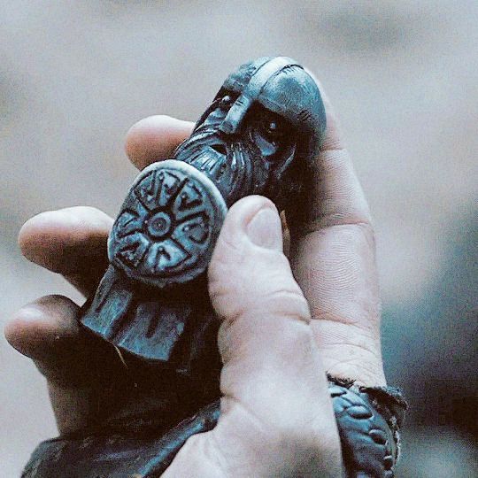 a person is holding a miniature carving of a viking figure with a shield in their hand. the carving shows a viking with a helmet, beard, and a round shield. the background is blurred, focusing attention on the figure and the person's hand.
