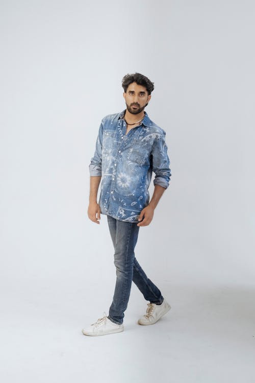 a fullbody studio shot of a young man walking. he's wearing a lightwash denim shirt with a unique pattern, dark wash jeans, and white sneakers. he has dark hair and a beard, and is looking directly at the camera. the background is plain white.