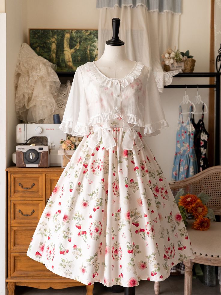 a white dress with pink strawberries printed all over it is displayed on a mannequin. the dress has a ruffled vneckline and a flowing skirt that reaches below the knee. a white blouse with puffy sleeves is worn underneath the dress. the background is a white room with wooden furniture and floral curtains.