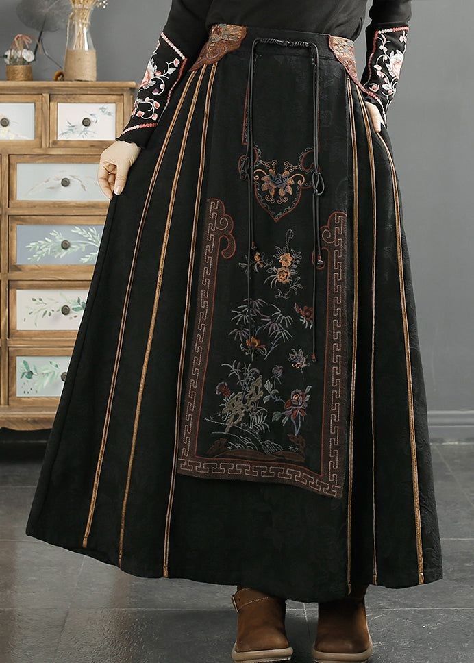 a closeup shot of a woman wearing a long, black, aline skirt with intricate floral embroidery. the skirt is made of a corduroylike material and has a high waist. the embroidery features a panel with a floral design, surrounded by a geometric border. the sleeves of the woman's top are also embroidered, and she is wearing brown boots. the background features a dark wooden dresser. the overall style is vintageinspired and bohemian, suggesting autumn or winter fashion.