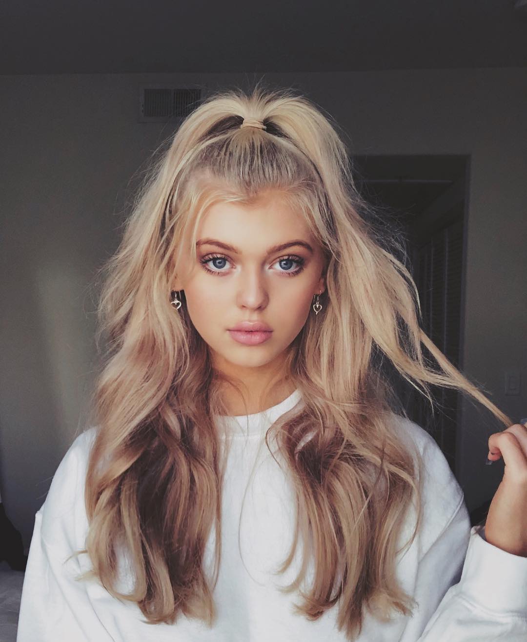 a portrait of loren gray with blonde hair in a half up half down ponytail hairstyle, wearing heartshaped earrings and a white sweater. she has blue eyes and a neutral facial expression. the background is a neutral gray color, likely a wall in an indoor setting.