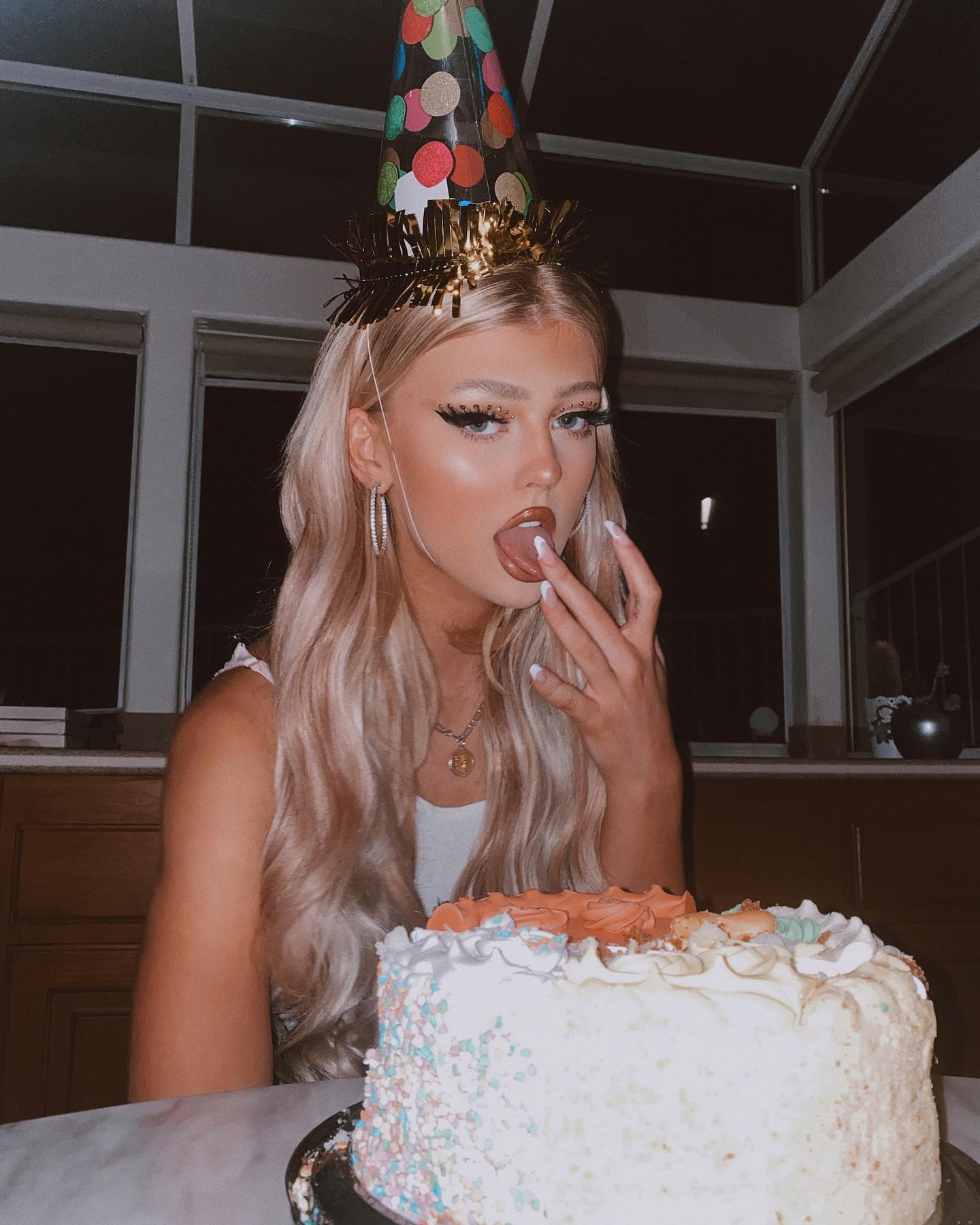 a woman wearing a birthday hat is sitting at a table with a cake. she has blonde hair, jewelry, and makeup. she is licking icing off her finger. the background shows a window and a wall.