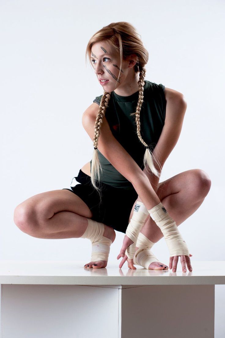 a young woman with blonde braids and athletic wear crouches on a white surface. her hands and feet are wrapped in white athletic tape, and she has black paint markings on her face, suggesting she's a fighter. the background is a plain white, indicative of a studio setting. the lighting is bright and even, illuminating the subject clearly.