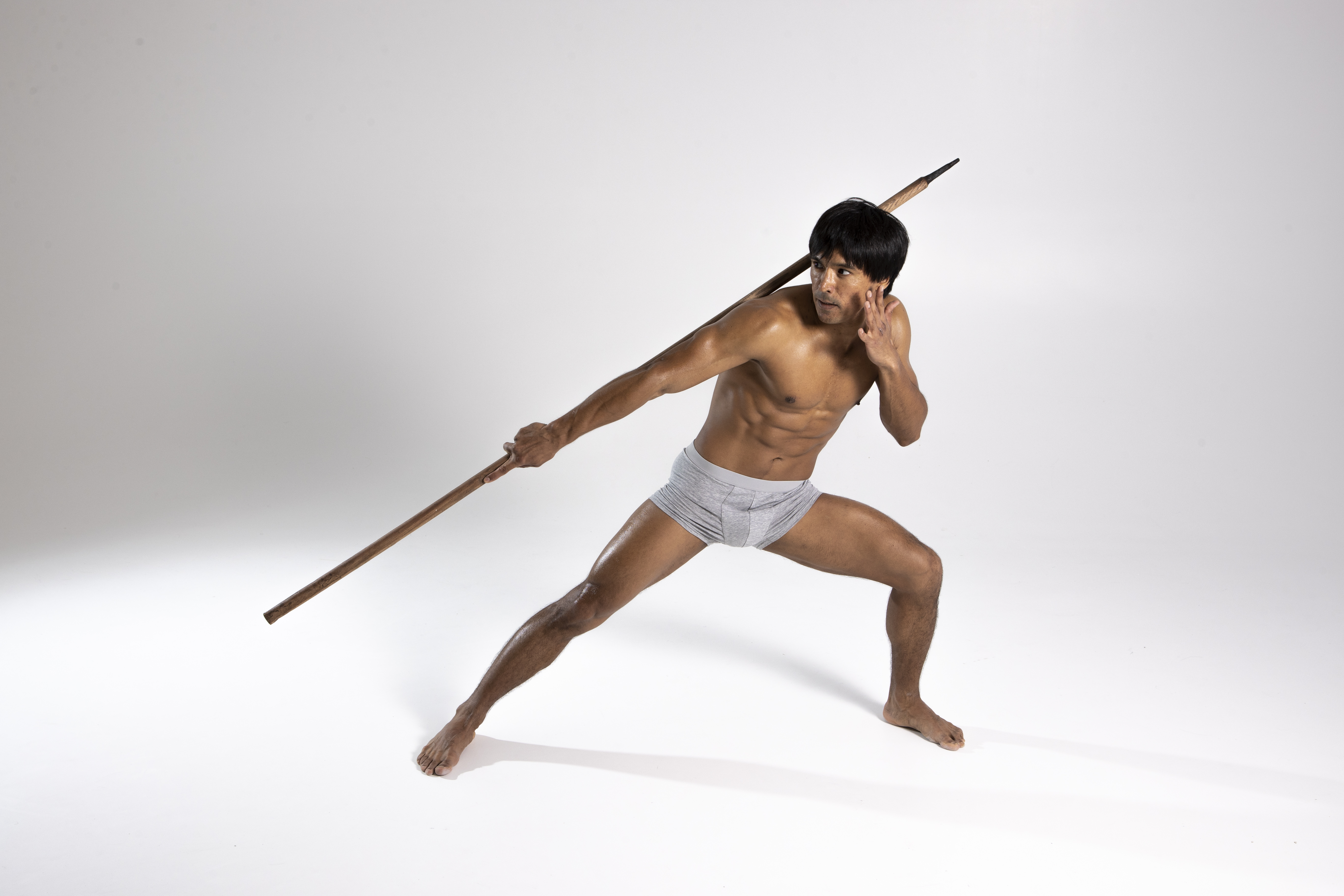 a shirtless man with dark hair and a muscular build is posing with a spear. he is wearing grey underwear and is barefoot. he is in a defensive posture, lunging forward with the spear extended and his other hand near his ear, as if listening for something. he appears to be alert and ready for combat, evoking a tribal warrior feel. the background is plain white.