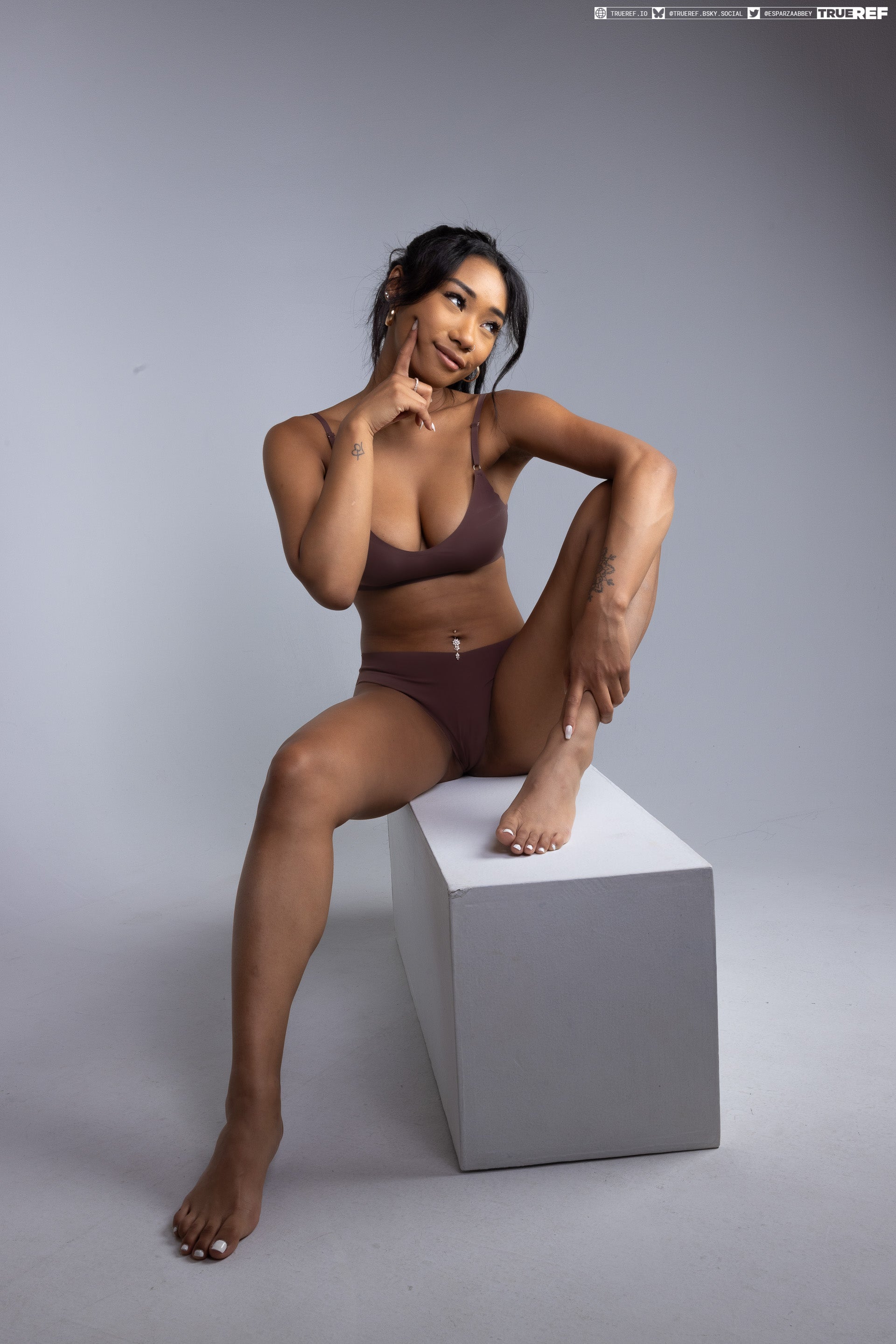 a tanned skin woman wearing a brown bra and underwear sitting on a white cube in a studio environment. she is looking thoughtful, with her index finger touching her chin. the background is a light grey. she has long hair, is wearing earrings and has a pierced navel. visible are also several tattoos on her arms and legs.