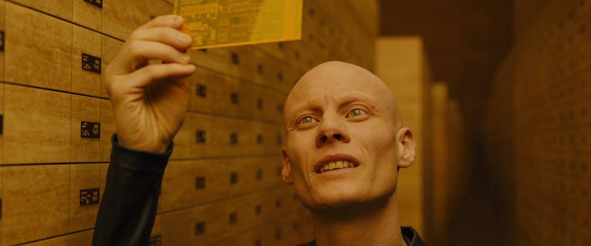 a bald man with striking pale skin and light eyes stands in a vault, surrounded by numerous goldcolored safety deposit boxes. he holds a translucent yellow card up, seemingly examining it with a focused, intrigued expression. the lighting is artificial and bright, casting a warm glow over the scene. the low camera angle adds to the sense of intrigue and mystery.