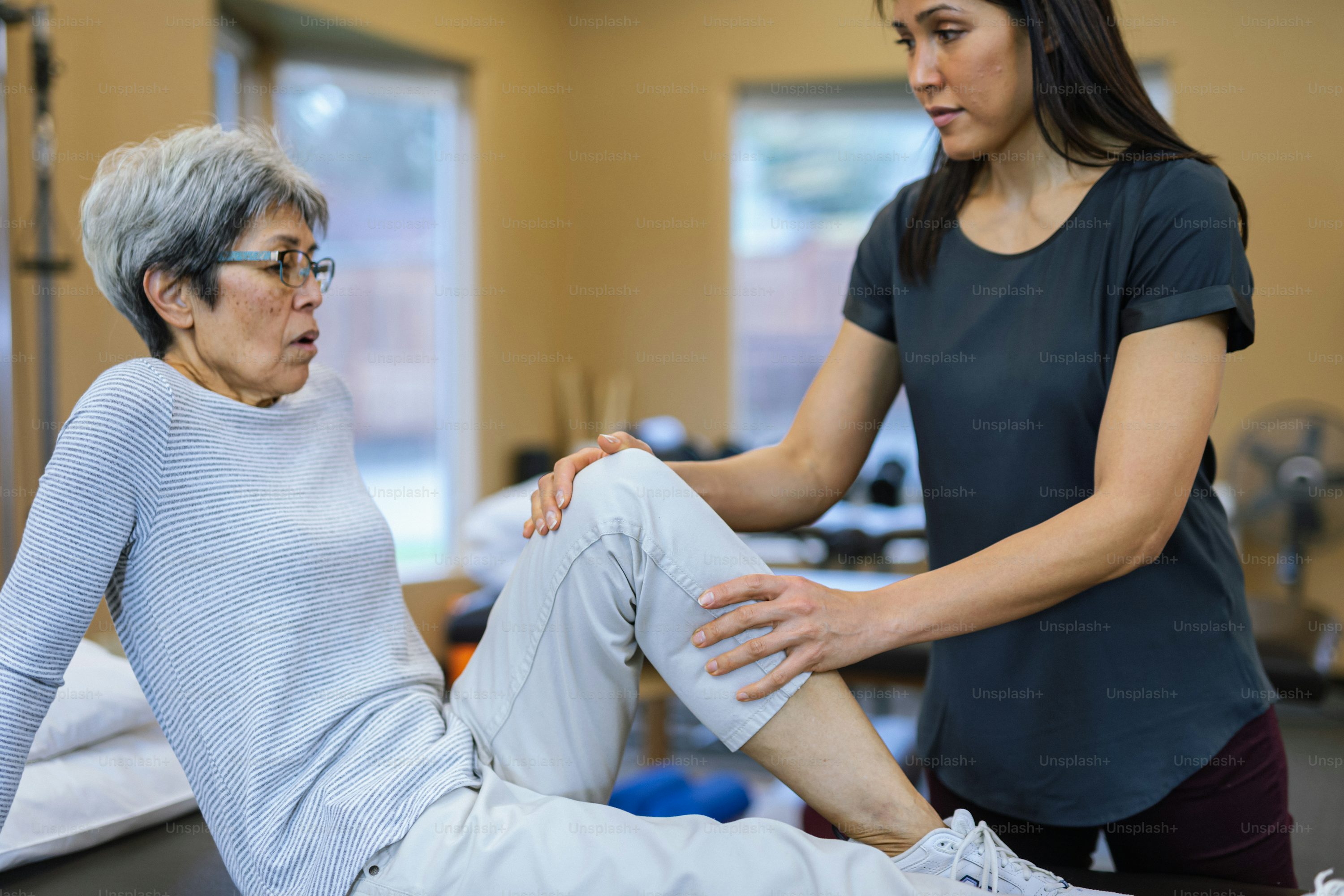 a woman wearing a grey longsleeve shirt with blue stripes and glasses is seated on a table with her knee up while a physical therapist wearing a darkgrey shirt holds her leg. they are in a room with windows and exercise equipment.