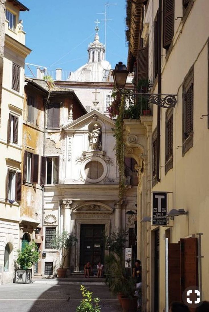 a narrow, sunlit alleyway in rome, italy, reveals the facade of a charming chapel with a statue of the virgin mary.  the chapel's architecture contrasts with the aging buildings that line the street.  in the distance, the dome of a larger church is visible. people are seated near the chapel's entrance.  the atmosphere is peaceful and evokes the historical charm of rome. a tobacco shop sign is visible on one of the buildings. plants and trees add to the picturesque scene.