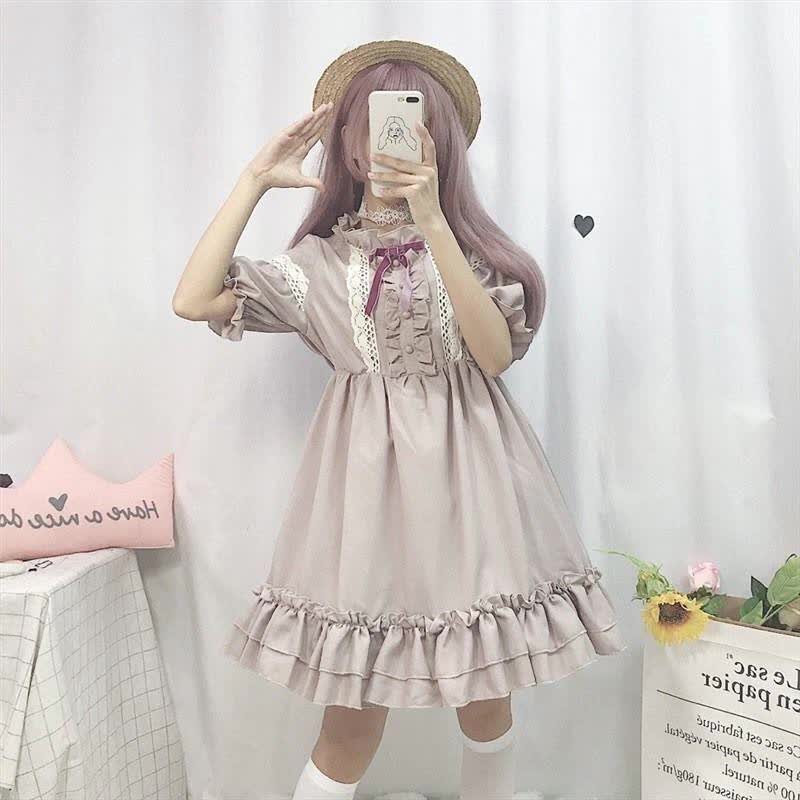 a young woman poses for a mirror selfie in a pastel purple lolita fashion dress. the dress has lace detailing, a ruffled skirt, and a bow at the neckline. she is also wearing a straw hat and white socks. the background is a white wall with a framed picture and a sunflower.