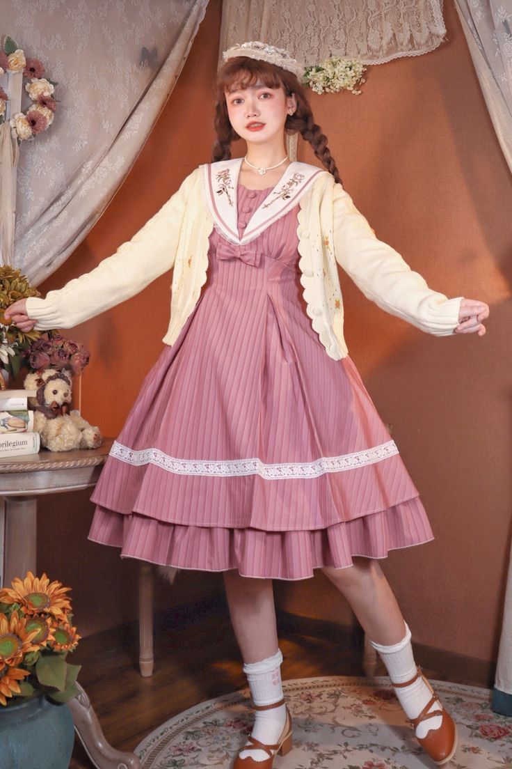 a young woman poses in a pink lolita fashion outfit. she wears a pink striped jumperskirt with lace trim, a cream cardigan with flower embroidery, and a white sailor collar with pink trim. she also wears a white headband and brown mary jane shoes. she is standing in front of a floral backdrop, and a teddy bear and book can be seen on a table to her left. the lighting is artificial and the perspective is eyelevel.