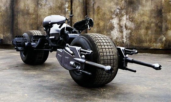 this is a picture of the batcycle from the batman movie franchise. the batcycle is a custommade motorcycle that is designed to be fast and powerful. it features a unique design with two wheels, a large, distinctive front wheel and a small back wheel. the batcycle is armed with guns and has a futuristic look that matches the batmobile. this is a dark grey batcycle with large black wheels and a sleek, black, curved seat. the batcycle is sitting on a concrete surface.  the back tire is partially obscured from view. the batcycle has a dark grey, matte finish with black accents and a silver gun muzzle.