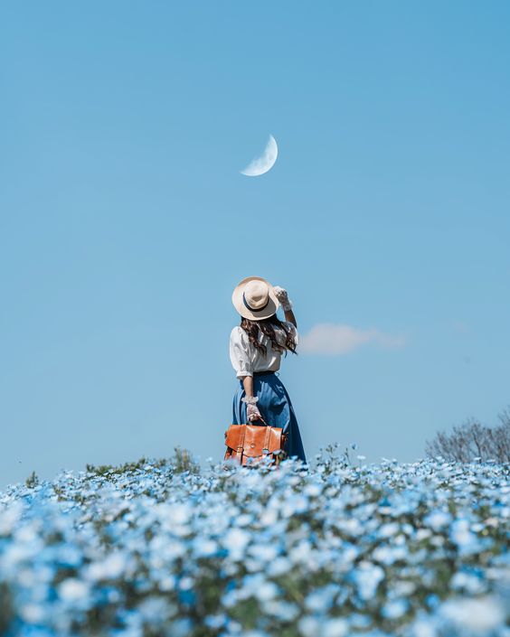 a woman in a white shirt and blue skirt, wearing a straw hat and holding a brown leather bag, stands in a field of blue flowers, gazing up at a crescent moon in a clear blue sky.