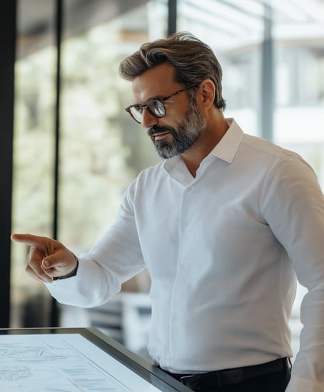 a middleaged man with graying hair and glasses is seen from the chest up, standing at a large, sleek touchscreen display. he's wearing a crisp white dress shirt and dark trousers. the man has a serious and focused expression as he interacts with the screen, his finger pointed toward it. he seems to be reviewing or presenting something. the background is blurred but suggests a modern, welllit office environment with large windows.  the overall tone is professional and sophisticated.