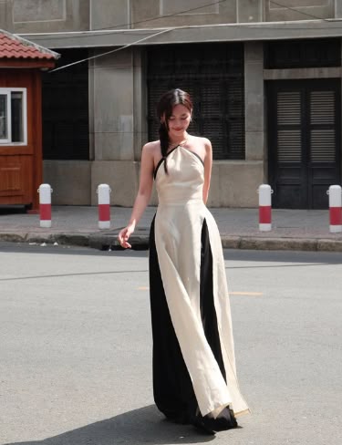 a woman is walking on a city street wearing a long, twotoned dress with a halter neckline. the dress has a lightcolored panel in the front and darker panels on the sides. she has her hair pulled back. the street is paved and surrounded by buildings with classical architectural details. there are red and white poles along the edge of the road.