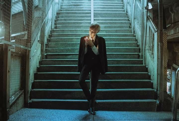 a man in a dark suit stands on a dimly lit staircase, holding a lighter and possibly smoking a cigarette. the atmosphere is moody and mysterious, with the man appearing pensive or contemplative. the setting is urban, possibly a city underpass or walkway at night. the image has a strong visual style with a focus on contrast, shadows, and atmosphere.