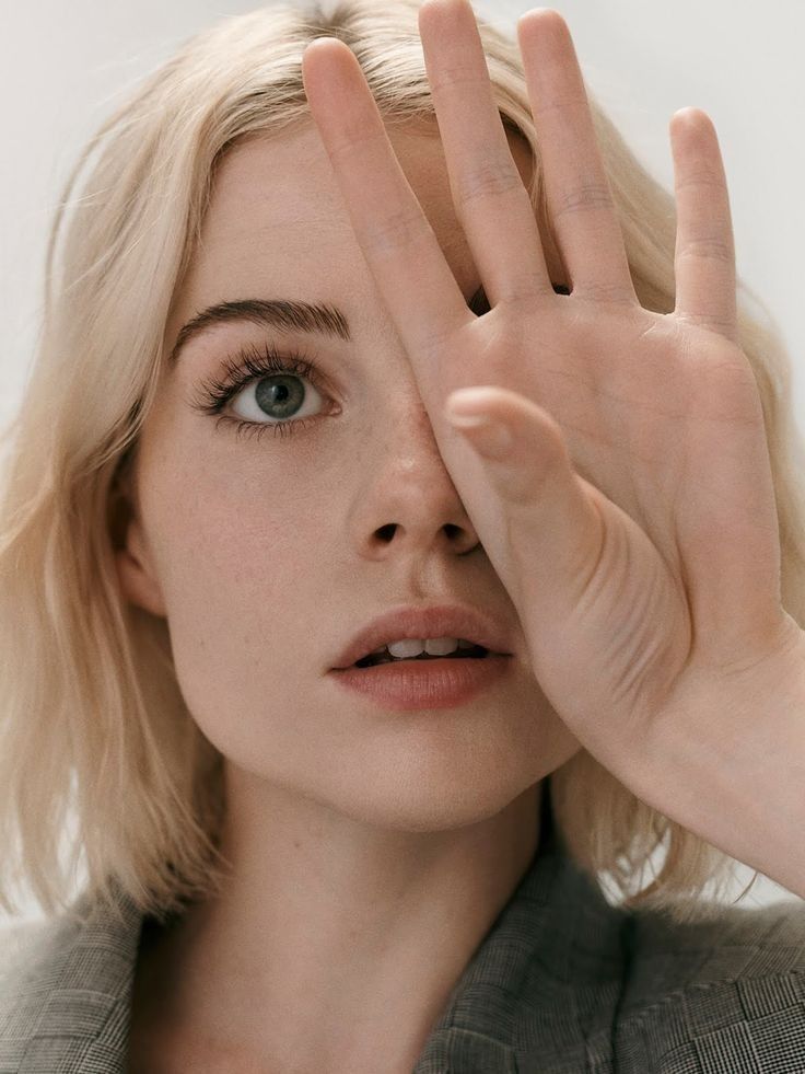 a blonde woman with short hair is covering one of her eyes with her hand. she is looking away from the camera. she is wearing makeup and her skin is clear. her lips are slightly parted.