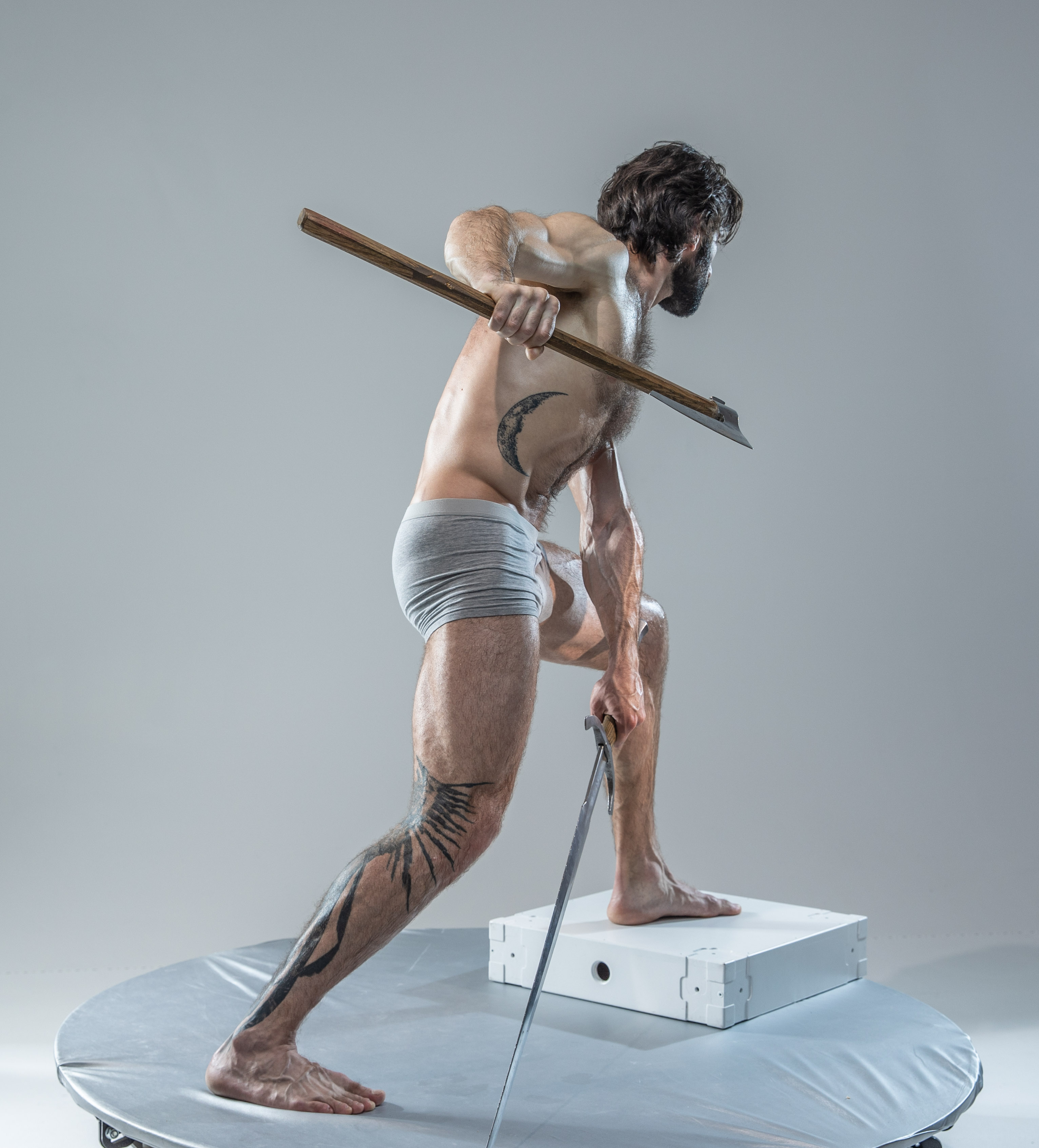 a muscular man with tattoos is captured in a dynamic pose against a grey studio backdrop. he grips a wooden axe, his body tensed as if ready to strike. the soft lighting emphasizes his physique and the raw energy of the scene. he stands on a small white platform, which is placed on top of a larger circular platform.  this setup, combined with the low camera angle, creates a sense of power and intensity.