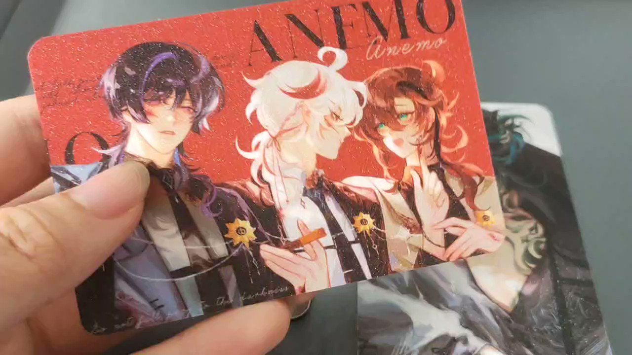 a closeup shot of a hand holding a textured, glittercovered card featuring animestyle fanart. the card depicts three characters: two boys and one girl, with a red background and the word "anemo" written across the top.  the word "anemone" is written smaller beneath it. the style of art is detailed and expressive. a portion of another piece of artwork is visible in the bottom right corner, suggesting a collection or set.