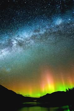 this image shows a stunning view of the night sky with the milky way and aurora borealis visible. the sky is a beautiful mix of green, yellow, pink, and purple, while the mountains in the foreground are silhouetted against the vibrant lights. the reflection of the aurora in the water adds to the magical ambiance of this scene.