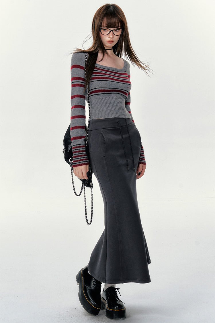 a fulllength studio shot of a young woman posing. she is wearing a grey and burgundy striped longsleeved sweater, a grey flare skirt, black platform shoes and eyeglasses. she is carrying a black shoulder bag.  the backdrop is plain white. the overall style is elegant and trendy.