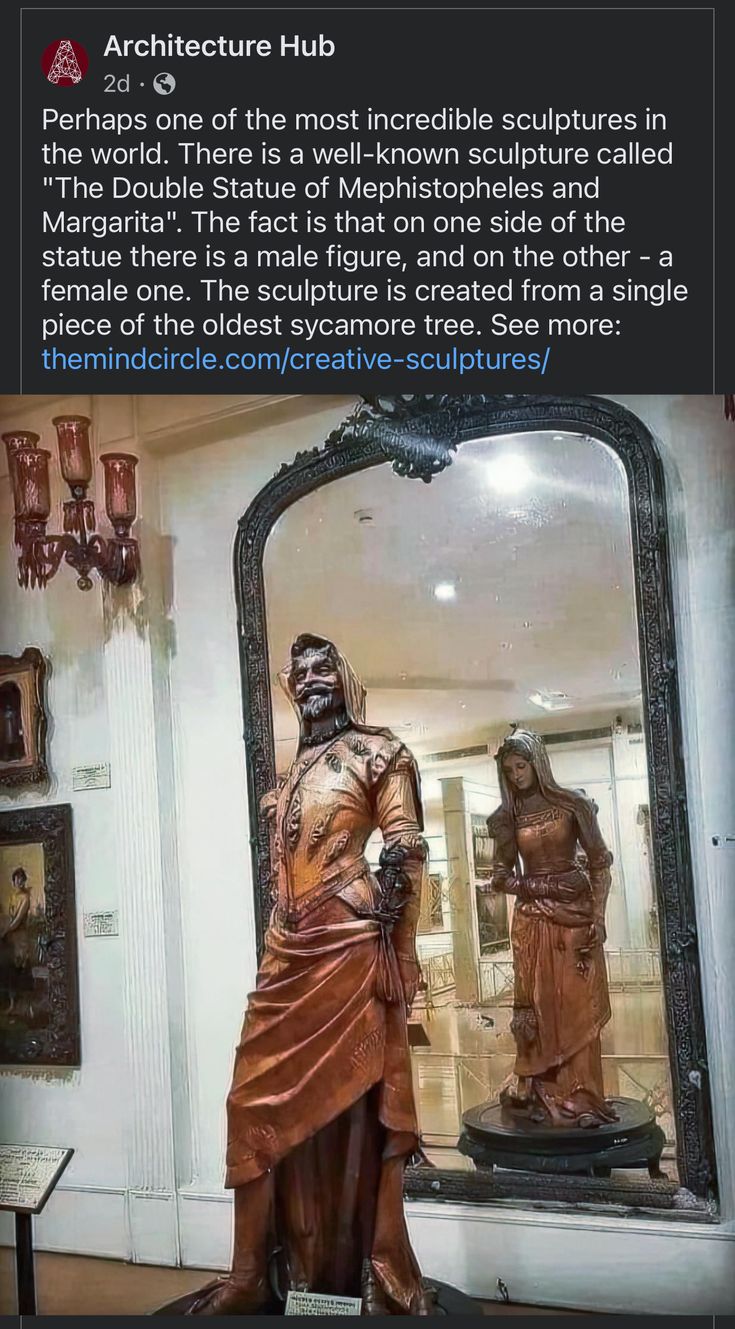 the image shows a facebook post from "architecture hub" featuring a wellknown sculpture called "the double statue of mephistopheles and margarita." the post highlights that one side of the statue depicts a male figure and the other a female figure, all carved from a single piece of sycamore tree. the image itself is a photo of the statue displayed in a museum setting, with a large mirror behind it, reflecting the female figure. other artwork and decorative elements are visible in the background.