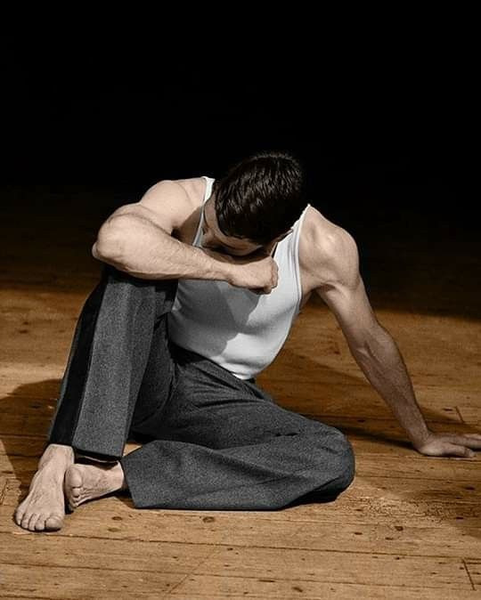 a man sits with his legs crossed on a wooden floor, with his head down and his elbows on his knees. he is wearing a white tank top and gray pants. he has a thoughtful expression on his face.  he is alone and seems to be deep in thought.