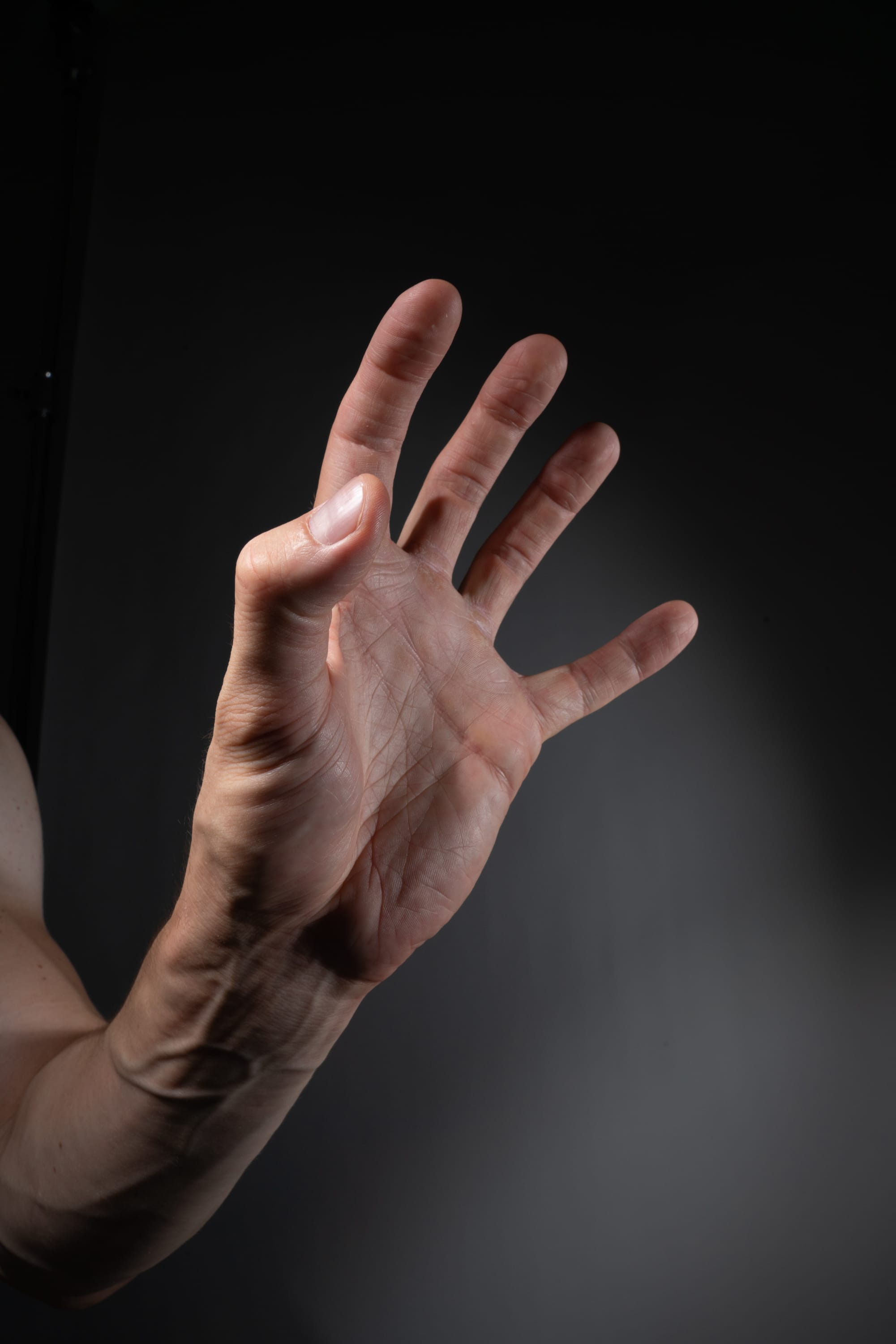 the image showcases a closeup of a man's left hand extended against a dark gray background.  the studio lighting highlights the details and textures of the skin and the veins along the arm. the hand is slightly curled inwards, with the thumb tucked close to the palm. the overall tone of the image is dark and dramatic, drawing attention to the form and gesture of the hand.