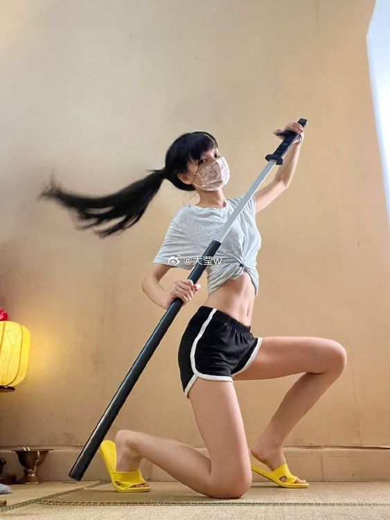 a young woman in athletic wear and a face mask strikes a dynamic pose with a sword indoors. the low camera angle emphasizes her athletic build and the sword's length. soft natural light illuminates the scene.