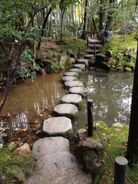 the image shows a tranquil stone path winding its way through a serene pond in a lush green forest. the pathway is made up of large, flat stepping stones, each carefully placed to create a harmonious flow.  the water is still and reflective, mirroring the surrounding trees and foliage. moss covers the ground and trees, adding to the serene atmosphere. sunlight filters through the trees, casting dappled light on the scene.