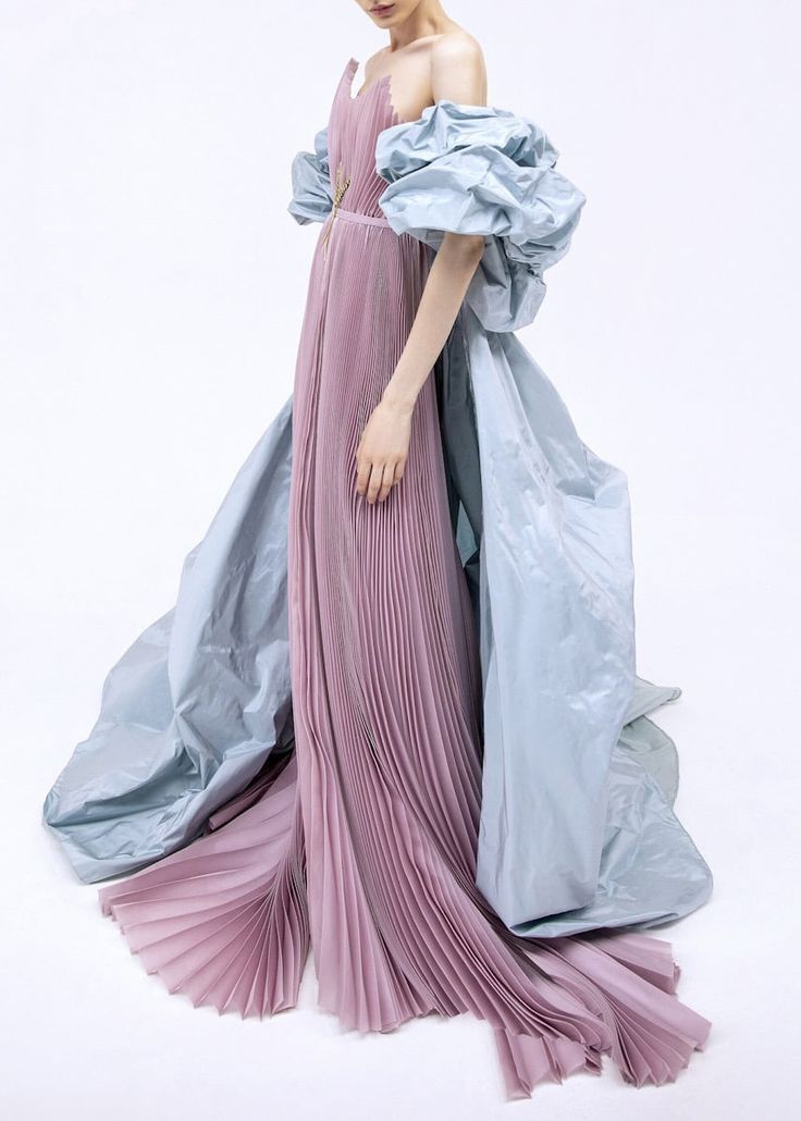 a woman is standing in a white room, wearing a long, elegant dress. the dress is pink and pleated with an offtheshoulder design. it features large, puffy blue sleeves and a flowing blue overskirt. the outfit creates a striking fashion statement.
