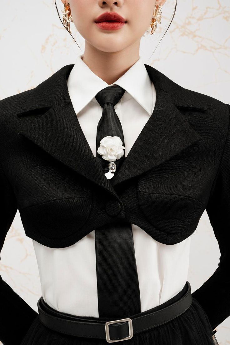 a woman wears a black cropped blazer, white shirt, and black tie with a white flower. she has a black belt with a silver buckle. the woman is wearing gold earrings.