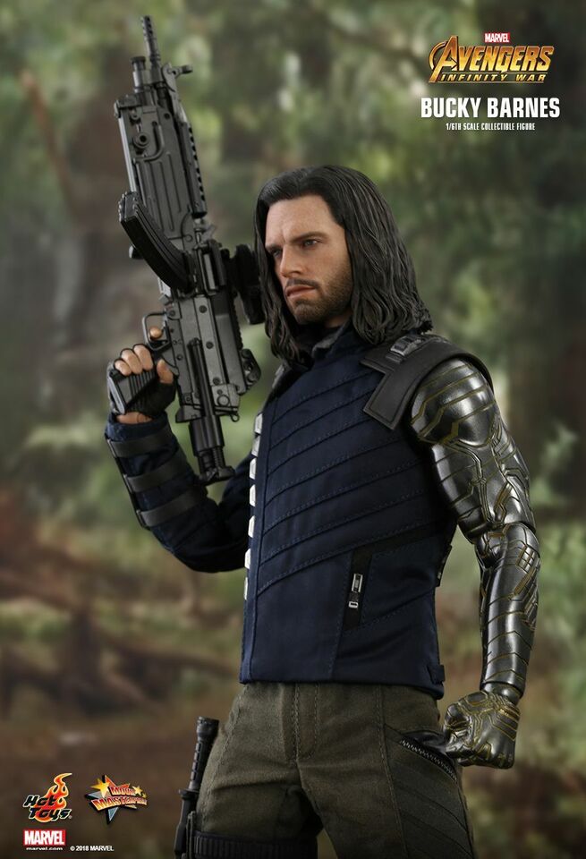 a closeup shot of a hot toys 1/6 scale collectible figure of bucky barnes from avengers: infinity war.  the figure is highly detailed, showcasing bucky's signature long hair, dark vest, and metallic prosthetic arm. he is holding a large rifle and is positioned against a blurred background of a forest.  the image includes hot toys and marvel logos.