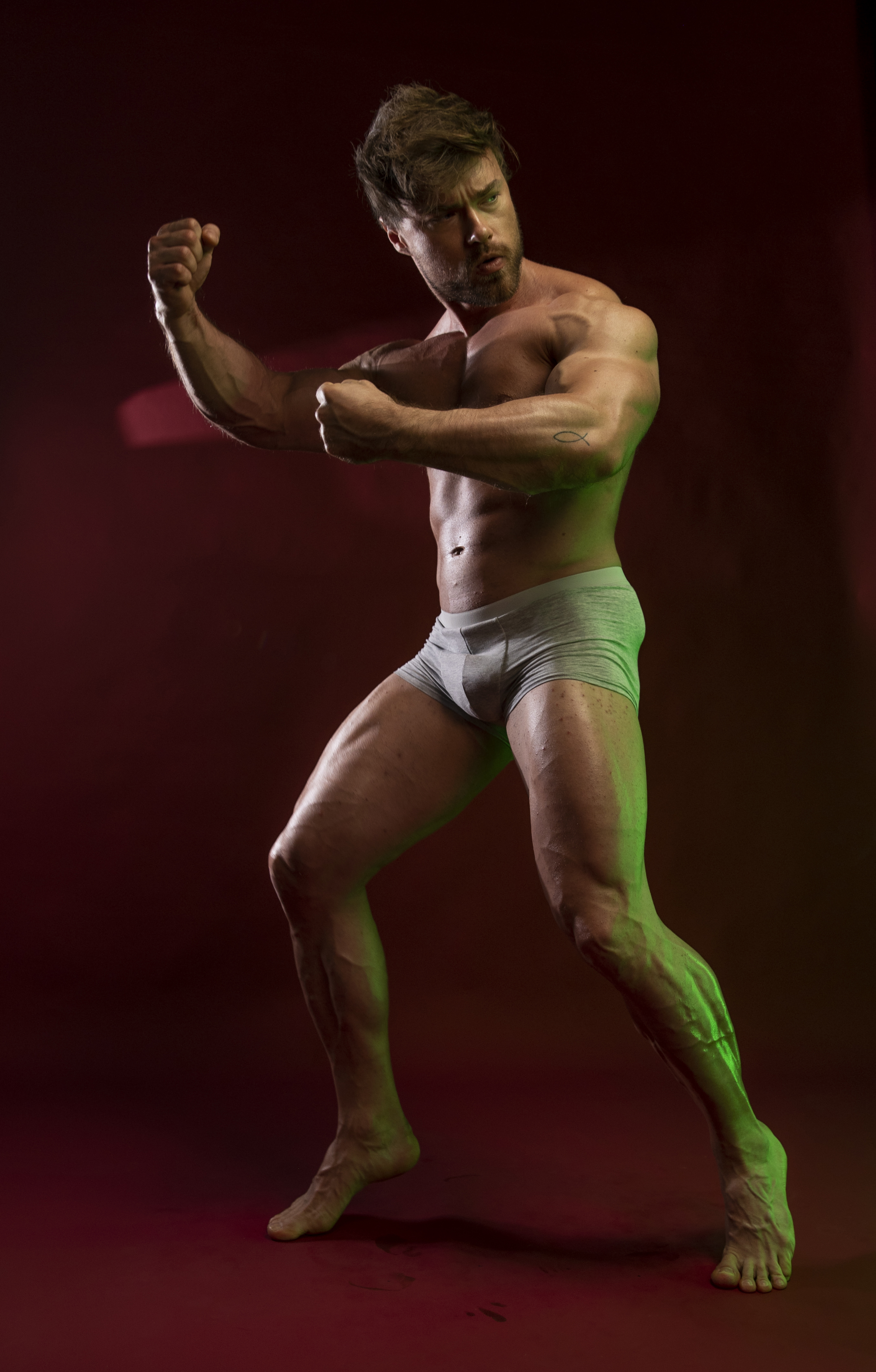 a muscular man wearing underwear is posing in a studio against a red background. he is barefoot and flexing his biceps and chest. he is looking to the side with a determined expression. the lighting creates a dramatic effect. the image highlights his physique.