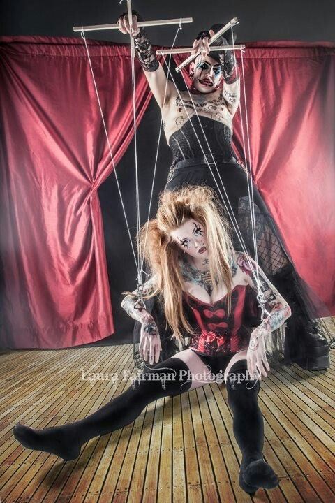 two women dressed in dramatic costumes are posing in a theatrical setting. one woman, the puppeteer, is wearing a black corset and a red bow, and is holding strings attached to a puppet. the second woman, the puppet, is wearing a red corset, black stockings, and a bow in her hair. they are both wearing a dramatic dark eye makeup. there is a red curtain and stage lighting in the background. the image is a dramatic and alluring depiction of a puppet show.