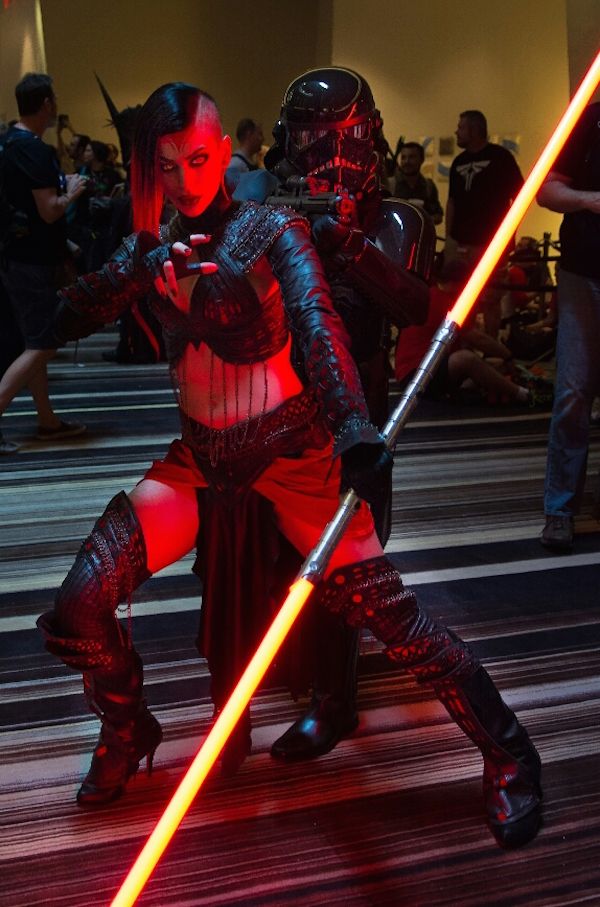 a woman dressed in a cosplay of a darth vaderlike character with a red lightsaber, surrounded by other people at a cosplay convention.