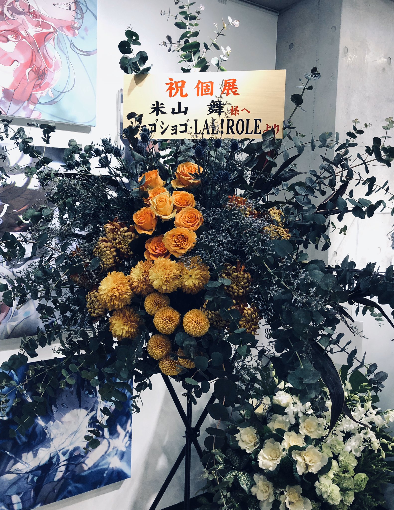 a vibrant floral arrangement stands in front of two framed artworks. the arrangement, primarily composed of orange and yellow flowers with green eucalyptus accents, is placed on a black stand.  a sign with japanese text is displayed prominently within the flowers. the overall ambiance suggests a celebratory occasion, perhaps an art exhibition opening or a similar event.