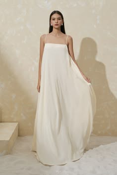 a woman in a simple, long white dress stands in a studio setting, looking at the camera with a relaxed and elegant pose. the dress is flowing and has a minimalist design, highlighting the model's beauty. the background is a simple, light beige, allowing the focus to remain on the woman and her outfit.