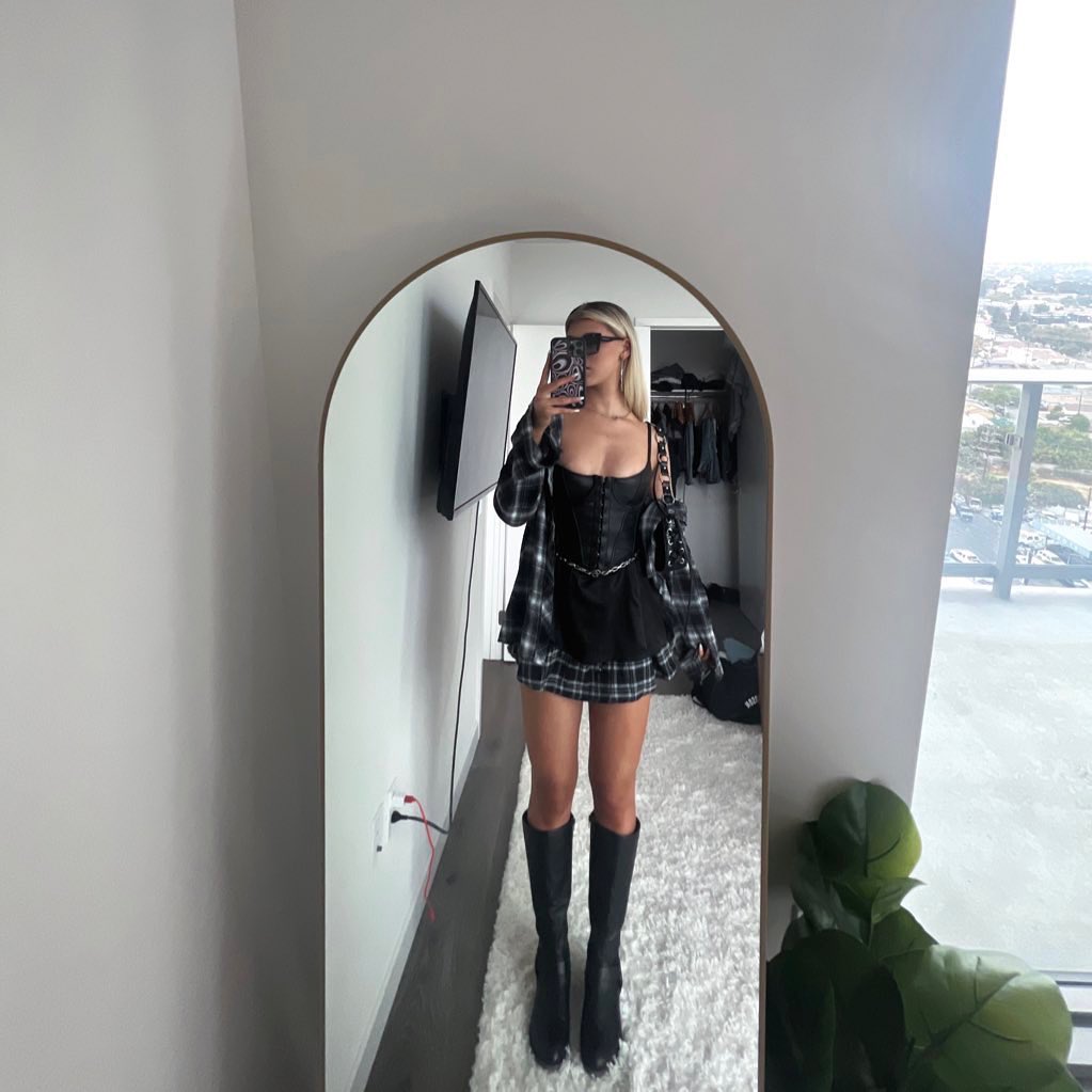 a woman is standing in front of a mirror in an apartment. she is wearing a black corset dress with a plaid mini skirt, kneehigh black boots, and a black and white plaid shirt. she is also wearing sunglasses and holding a phone. behind her is a window, closet, and a plant. she is standing on a white rug.