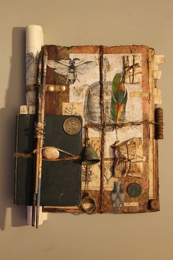 the image shows a piece of mixed media assemblage art. it appears to be constructed around a vintage book or journal as its base. the artwork incorporates various found objects and ephemera tied together with twine. there are paintbrushes strapped to the side of the book, along with an antique coin, a small bell, and a feather. the surfaces are covered with layers of old paper, botanical drawings, entomological illustrations (particularly a wasp), and scientific diagrams. the overall composition has a vintage, naturethemed aesthetic with a textured, handmade quality.